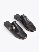 Mocasín tipo slide color marrón oscuro con efecto gastado, confeccionado en cuero vacuno. Presenta detalles de picado en la capellada y laterales, forrería de cuero vacuno, hebilla y puntera metálica.