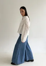 Blusa de brodery color blanco, con cuello redondo, mangas largas abullonadas con puños elásticos y cierre frontal con botones forrados. Presenta detalles de puntillas verticales en el frente.