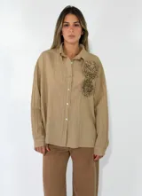 Camisa de corte oversize color beige, con cuello clásico, cierre frontal de botones y detalle de flores de tela aplicadas en el pecho.