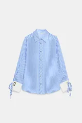 Camisa de cuello solapa y manga larga con rayas verticales celestes y blancas. Cuenta con puños anchos blancos contrastantes, ajustables con hebilla dorada y lazos, y cierre frontal con botones dorados.