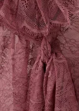 Blusa de encaje color bordó con cuello gorguera, lazo frontal y mangas largas con volantes en los puños. Presenta un diseño recto, cierre de cremallera en la parte posterior y forro interior.