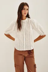 Camisa de algodón color celeste con cuello alto con volados, mangas tres cuartos abullonadas y cierre frontal con botones. Presenta un diseño con dos franjas verticales de bordado intrincado en color negro en el frente.
