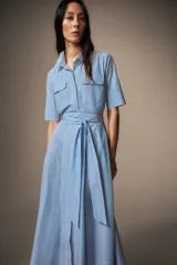 Vestido midi de algodón con estampado de rayas verticales, cuello solapa, manga corta con puños y cierre frontal de botones. Presenta bolsillos con solapa en el pecho y un cinturón de la misma tela para ajustar la cintura.