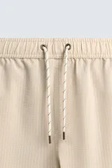 Bañador largo de hombre color rosa pálido con textura seersucker. Presenta cintura elástica ajustable con cordón blanco, bolsillos laterales y un bolsillo trasero de plastrón.