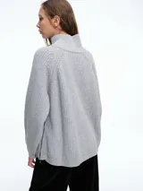 Sweater de tejido de punto acanalado en color blanco, con cuello alto y cierre metálico diagonal en el hombro. Presenta mangas largas con diseño raglán y corte holgado.