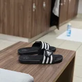 Sandalias tipo slide Adidas Adissage, color negro con tres franjas blancas en la tira superior ajustable con velcro. La plantilla presenta un diseño texturizado con protuberancias para masaje.
