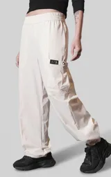 Pantalón cargo de corte oversize color beige, confeccionado en microfibra técnica. Presenta cintura elástica, bolsillos laterales y un bolsillo cargo con cierre de broche snap en el muslo. Incluye un sistema de elásticos con tancas en los tobillos para regular la apertura.
