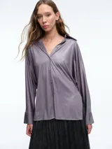 Camisa de corte oversize confeccionada en tejido jersey brillante con caída fluida. Presenta cuello en punta, cierre frontal con botones, mangas largas con puños anchos y tajos laterales en el ruedo.