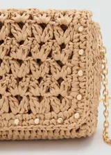 Bolso tipo bandolera de tamaño mini, confeccionado en fibras naturales tejidas con diseño de espiga. Presenta cierre de solapa con botón magnético, detalles de tachas metálicas en los bordes, forro interior y asa larga de cadena metálica dorada.
