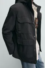 Parka técnica de corte relajado con interior ligeramente acolchado. Presenta cuello subido con capucha ajustable, cierre frontal con cremallera oculta por solapa con botones a presión, y cuatro bolsillos frontales de plastrón con solapa. Incluye cintura ajustable con cordón interior, puños con rib y aberturas laterales con cremallera en el bajo.