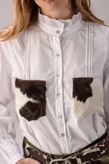 Camisa blanca de manga larga con cuello alto gorguera con volados y detalles de alforzas verticales en el frente. Cuenta con dos bolsillos en el pecho de pelo con estampado de vaca en tonos marrones y blancos.