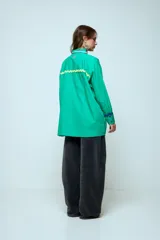 Camisa azul de corte oversize, con cierre frontal de botones y detalles de cintas onduladas en colores contrastantes a lo largo de la botonadura y en los puños. Presenta cuello clásico y mangas largas.