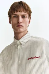 Camisa de corte holgado en mezcla de algodón Oxford, con diseño a rayas, cuello americano y cierre frontal de botones. Cuenta con un bolsillo superior en el pecho con texto bordado en rojo, mangas largas con puños abotonados y bajo recto.
