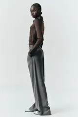 Pantalón gris de tiro alto con bolsillos delanteros y traseros, detalle de pliegues frontales y cierre frontal con cremallera y botón.