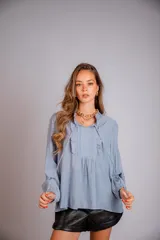 Camisa de color celeste con volados decorativos en el cuello y la parte delantera, cuenta con cordones para ajustar el escote, mangas largas con puños y corte holgado.