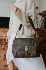 Cartera de hombro con textura efecto cocodrilo en color marrón oscuro. Presenta un diseño rectangular con solapa frontal, cierre metálico a presión y una correa de cadena gruesa en tono dorado.