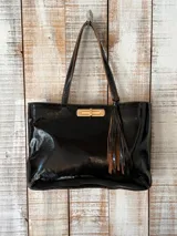 Cartera tipo tote de cuero negro con acabado brillante, cierre frontal metálico dorado y detalle de flecos colgantes en el lateral. Cuenta con doble asa de hombro.