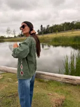 Campera de jean color verde oliva, con diseño de figuras geométricas en terciopelo aplicadas en la espalda y mangas, bordadas a mano con costuras decorativas. Presenta un corte holgado y etiqueta de cuero en la parte superior de la espalda.