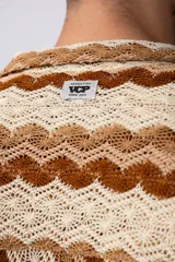Camisa de manga corta confeccionada en tejido de crochet con diseño de ondas verticales en tonos tierra y beige. Presenta cuello solapa y cierre frontal con botones.