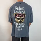 Remera de corte oversize color gris azulado, con estampa gráfica en la espalda que incluye un oso con anteojos y bandana, acompañada de la frase 'Richer, Louder & Unbothered Than Ever'.