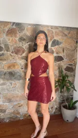 Vestido corto de lentejuelas color bordó, con escote halter y diseño cut-out en la cintura. Presenta breteles finos regulables que se cruzan en la espalda.