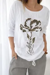 Remera blanca de línea italiana, confeccionada en algodón y viscosa, con calce holgado y cuello redondo. Presenta un estampado frontal de una flor en tonos oscuros con detalles dorados y la frase "Ladybee". Incluye tiras regulables en la parte inferior para ajustar el largo.