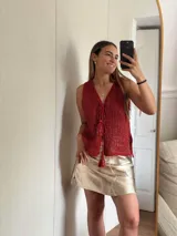 Chaleco tejido en punto calado con hilo de algodón y lurex, de color negro con destellos plateados. Presenta escote profundo en V, cierre frontal con cordones entrelazados terminados en borlas y un corte recto y holgado.