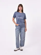 Remera de manga corta color beige claro con cuello redondo. Presenta un estampado gráfico central con un plato de pasta rodeado por un borde azul y blanco, sobre un fondo de franjas verticales rojas y blancas. El estampado incluye texto en cursiva en tonos turquesa y rojo.