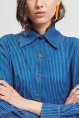 Camisa de corte holgado confeccionada en crepé fino con estampado de rayas verticales. Presenta cuello clásico de tamaño ligeramente ampliado, mangas con pinzas laterales y puños anchos.
