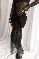 Vestido corto negro de encaje transparente, con mangas largas y falda ajustada con detalle lateral asimétrico tipo pareo.