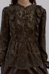 Blusa de manga larga con cuello redondo, confeccionada en tejido semitransparente con estampado paisley en tonos marrones oscuros. Presenta detalles de volantes y apliques de terciopelo en el frente y hombros, con cierre frontal de botones forrados.