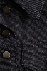 Chaleco corto de denim de algodón rígido, color azul oscuro, con cuello, cierre frontal de botones de metal y dos bolsillos superiores con solapa y botón. Presenta un corte ajustado con canesú y costuras visibles que aportan estructura.