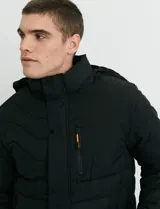 Campera puffer con capucha y cuello alto, de color negro, con cierre frontal y solapa con botones a presión, y un bolsillo vertical con cierre en el pecho.