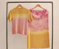 Conjunto de dos piezas compuesto por una remera de manga corta con cuello alto y una falda midi de corte recto. Ambas prendas están confeccionadas en lana merino con un diseño de teñido artesanal en degradé de tonos amarillos y rosados.