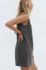 Vestido corto sin mangas, de corte recto, con cuello redondo y cierre frontal de botones. El tejido es tweed o similar, en tonos grises y negros jaspeados.