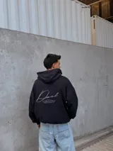 Canguro negro con capucha, corte oversize y diseño con hombros caídos. Presenta detalles bordados en la espalda con la palabra "Dual" en tipografía cursiva junto a texto descriptivo en tonos monocromáticos.