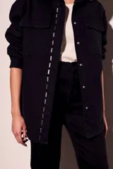 Sobrecamisa negra de algodón con corte oversize, cuello clásico, cierre frontal con botones a presión, dos bolsillos de parche en el pecho y apliques metálicos en cuello y solapa.