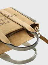 Cartera tipo tote de Marc Jacobs, confeccionada en una combinación de gamuza y cuero texturizado en tonos tierra. Presenta el logo 'THE TOTE BAG' estampado en el frente, doble asa superior y correa ajustable extraíble.