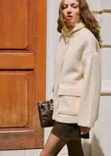 Chaqueta de corte recto y diseño medio, confeccionada en tejido bouclé color beige. Presenta cuello alto, manga larga, cierre frontal oculto con cremallera y botones a presión, y dos bolsillos de parche laterales.