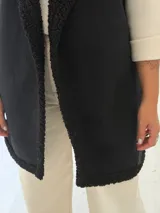 Chaleco largo de gamuza sintética color negro, con interior y bordes de corderito sintético al tono. Presenta un diseño abierto sin mangas, ideal para superponer sobre otras prendas.