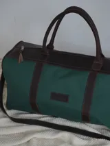Bolso tipo duffel de gran tamaño, confeccionado en lona color verde oliva con detalles y correas en cuero color suela. Cuenta con cierre superior y etiqueta de cuero con el logo de la marca.