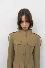 Blazer entallada color beige con cuello subido, manga larga con hombreras y trabillas en los hombros. Presenta bolsillos de plastrón en el delantero, detalle de solapas en el pecho y cierre frontal con botones a presión.