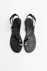 Sandalias planas de cuero color negro, con tiras finas, sujetador para el dedo y detalle de anilla metálica en la puntera. Cuenta con tira ajustable alrededor del tobillo con hebilla plateada.