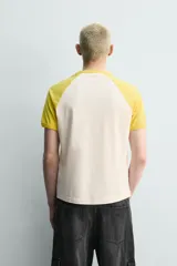 Camiseta de corte relajado con mangas raglán cortas en color amarillo y cuerpo en color crema. Presenta un estampado gráfico frontal con temática de campamento de básquetbol y un cocodrilo.