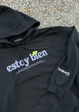 Canguro negro con capucha y cordones ajustables. Estampado frontal con la frase "estoy bien" en letras lilas y una flor verde.