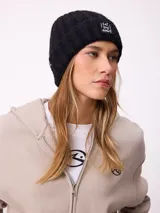 Gorro tipo beanie de punto acanalado en color lila, con parche rectangular negro en el frente que incluye la inscripción 'EAT THE WAVE'.