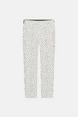 Pantalón blanco con estampado de lunares negros, de tiro medio con cintura elástica en la espalda, bolsillos delanteros y falsos bolsillos de vivo en la espalda. Bajo acabado con aberturas laterales y cierre frontal con cremallera, botón interior y gancho.