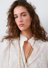 Blusa blanca de algodón con bordados florales, cuello alto con volados y mangas largas abullonadas con puños ajustados.