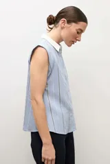 Camisa sin mangas celeste con rayas verticales blancas y finas líneas oscuras, con cuello camisero blanco y cierre frontal de botones.