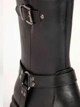 Bota alta negra de estilo rocker, con suela track gruesa, hebilla decorativa en la parte superior y tiras con tachas y argolla metálica en el tobillo.